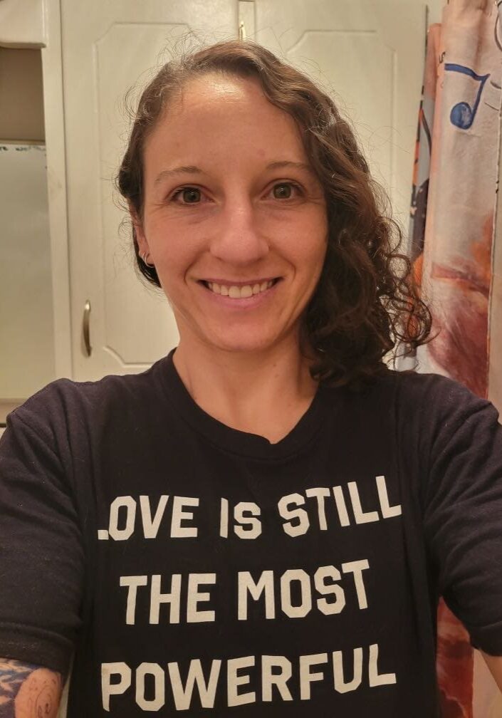 Michaela, a woman between the age of 25-35. Long, curly brown hair. Smiling. Has a tshirt saying "love is still the most powerful: