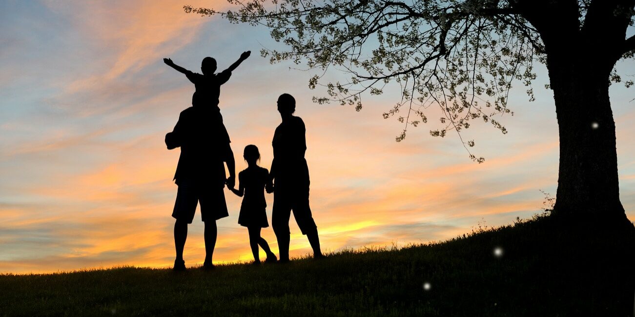 A sunset photo of a family of four. Father and mother holding their daughter's hand in between them and the son is on the father's shoulders with arms wide opening looking at the sunset.